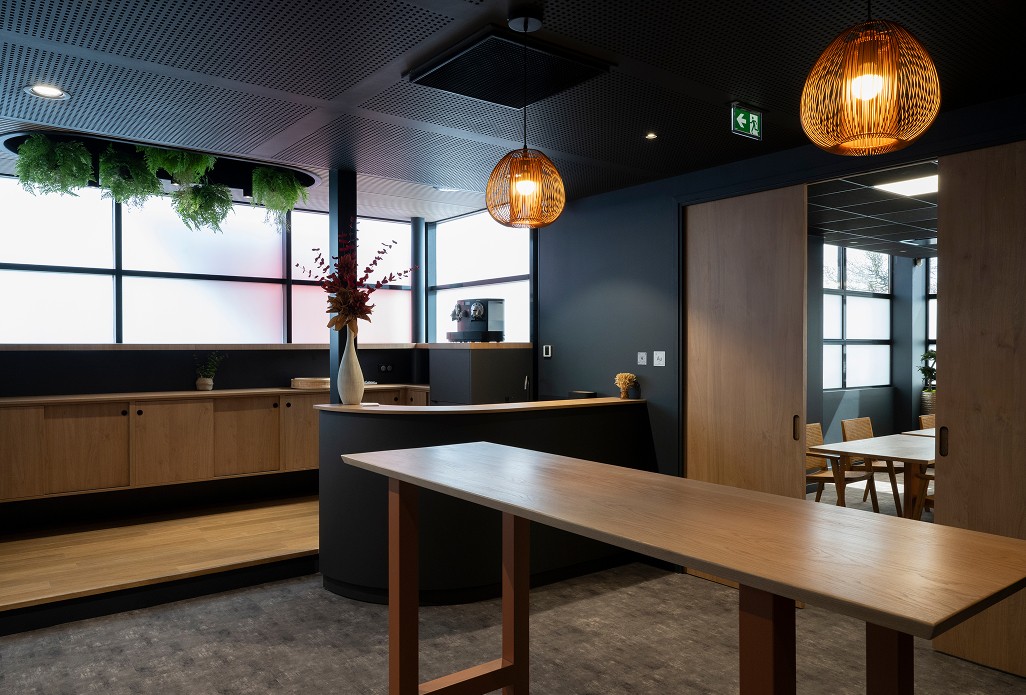 Vue d'un espace d'accueil moderne avec comptoir noir mat, meubles en bois clair, suspensions en rotin et lumière naturelle filtrée – ambiance chaleureuse et professionnelle.