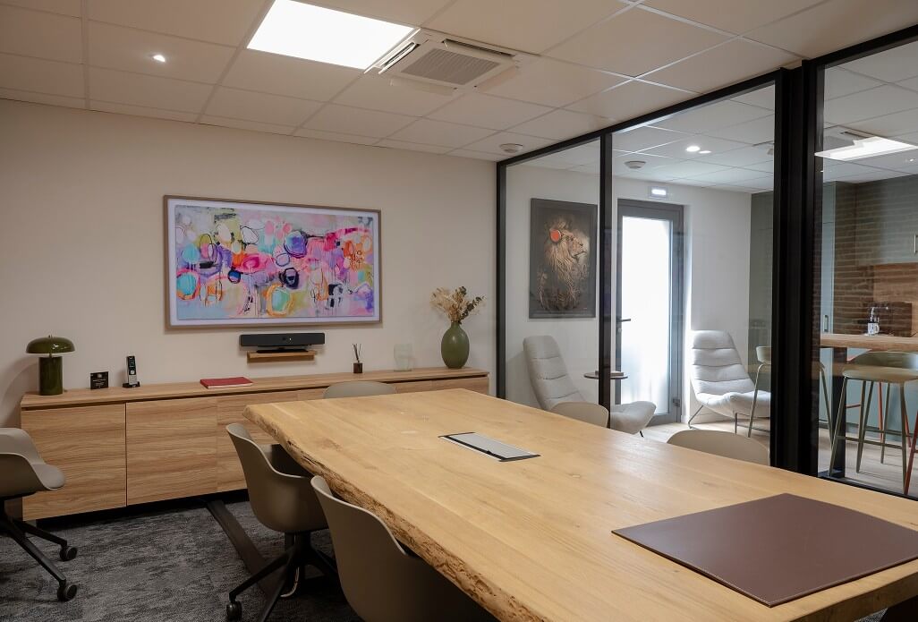 Salle de réunion contemporaine avec grande table en bois brut, chaises design, décoration murale artistique et cloison vitrée donnant sur un espace détente.